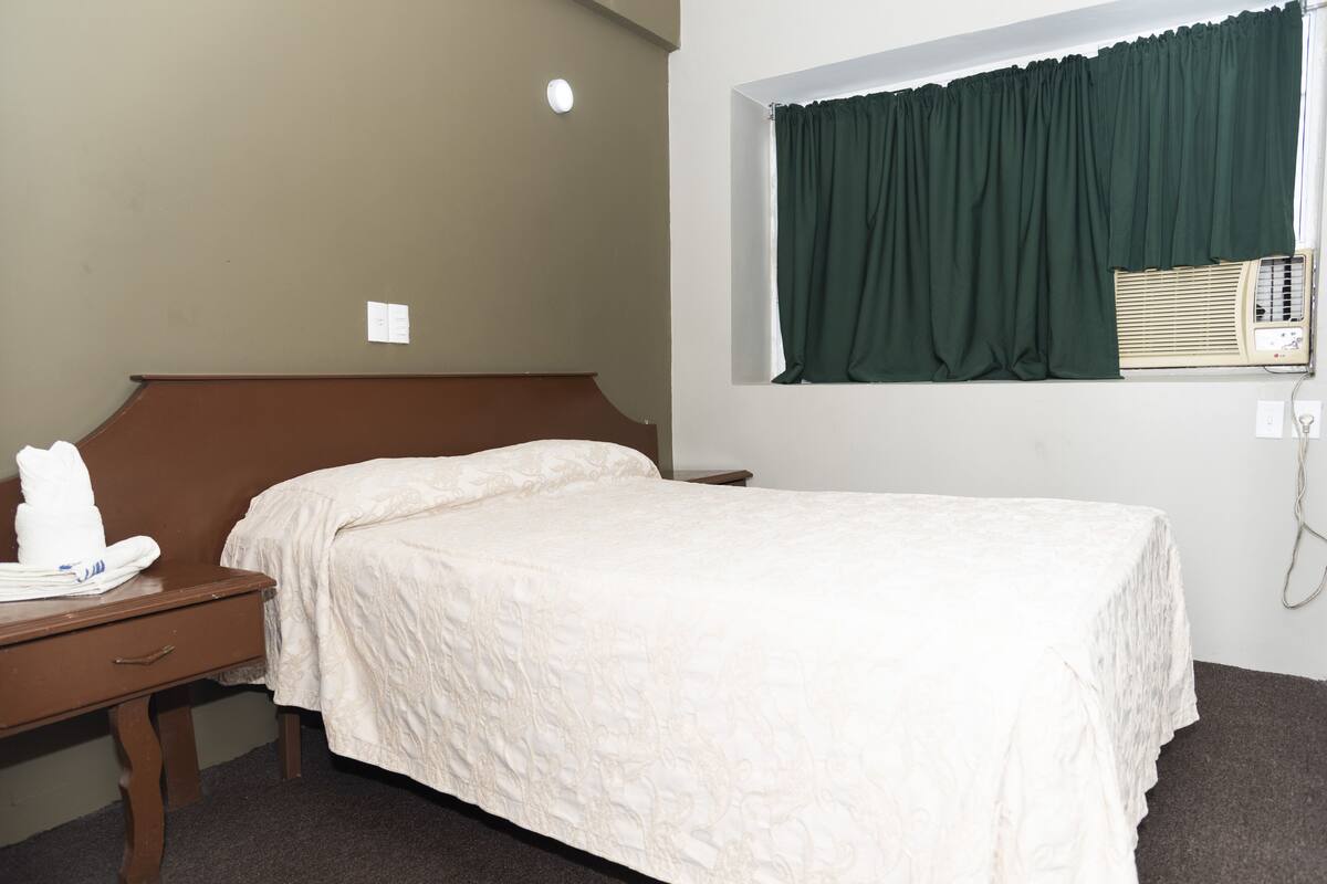 A hotel room with a single bed, a wooden nightstand, a desk, and a window with a green curtain.