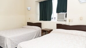 Habitación de hotel con dos camas individuales, una mesita de noche de madera, una ventana con cortinas verdes y un equipo de aire acondicionado.