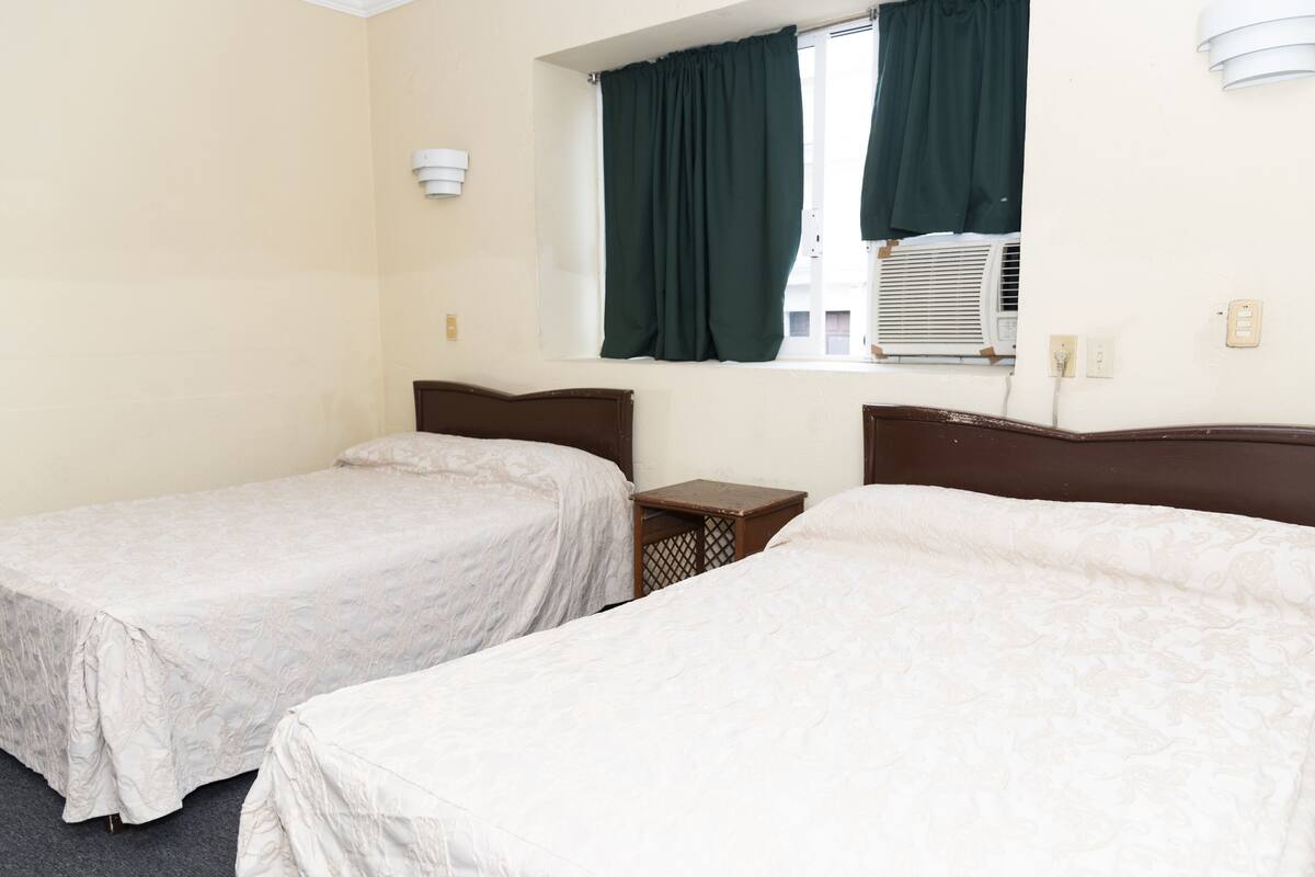 A hotel room with two single beds, a small wooden nightstand, a window with green curtains, and an air conditioning unit.