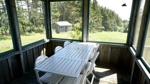 Un espacio al aire libre cubierto con una mesa y sillas de madera, y vista a una casa y un bosque al fondo.