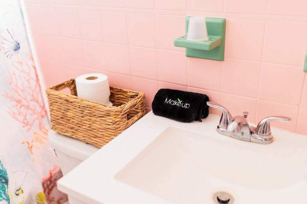 Ein Badezimmer mit einem weißen Waschbecken, einer grünen Seifenschale, einem schwarzen Handtuch mit der Aufschrift 'Makeup' und einem Korbwäscher mit einer Toilettenpapierrolle.