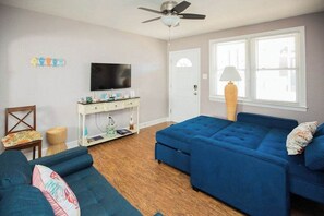 A living room with a blue sofa, a television, a ceiling fan, and a window with blinds.