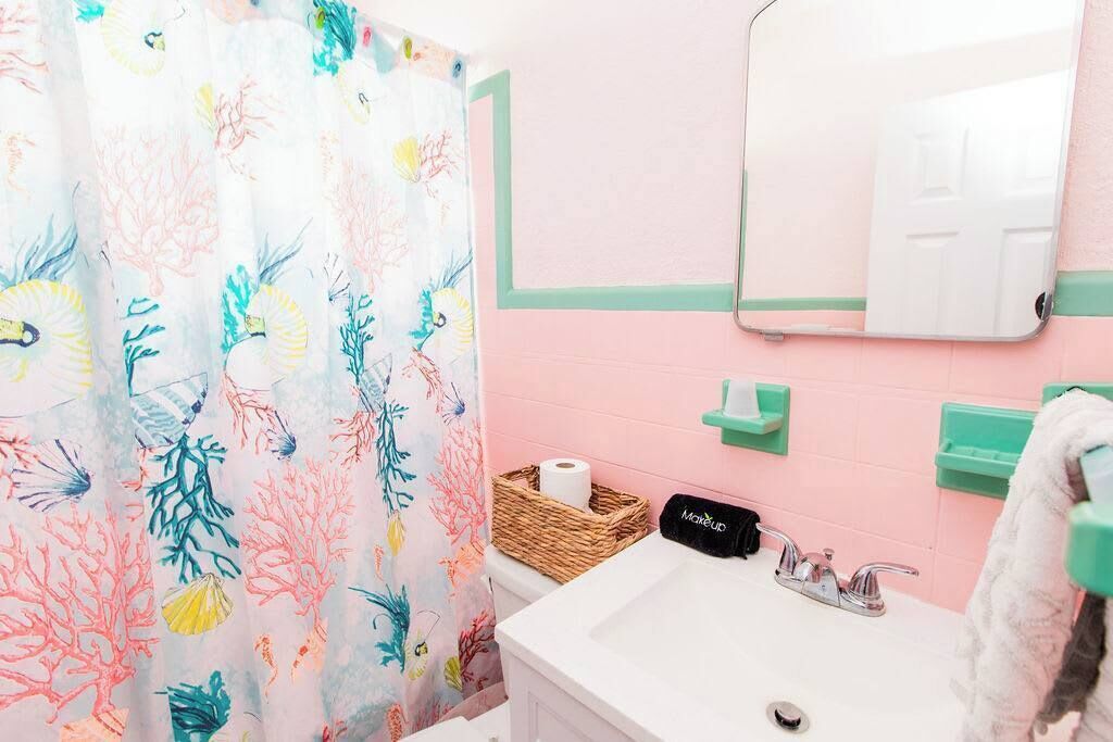 A bathroom with a pink wall, a white sink, a mirror, a green towel rack, and a shower curtain with a coral pattern.