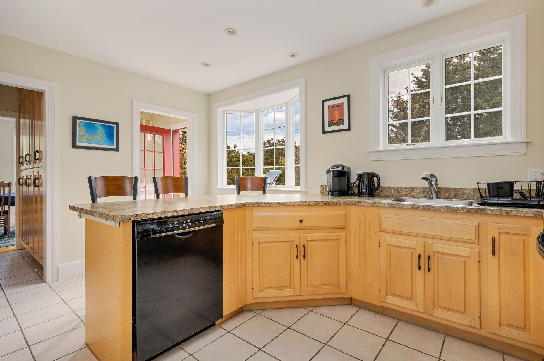 A kitchen with a dishwasher, wooden cabinets, a granite countertop, a coffee maker, and a window with a view of trees.