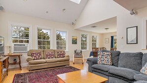 A spacious living room with a high ceiling, large windows, a wooden coffee table, and a patterned rug.
