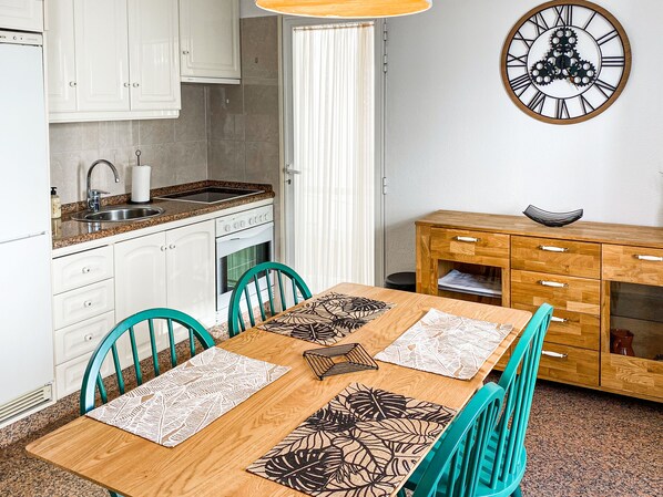 Une cuisine avec des meubles blancs, un évier et une cuisinière. On y trouve une table à manger en bois, des chaises et une horloge murale.