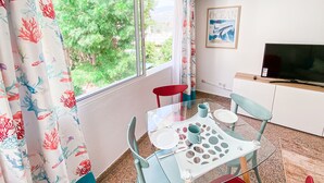 Un coin repas avec une table en verre, deux chaises et une chaise rouge. Un téléviseur est fixé au mur.