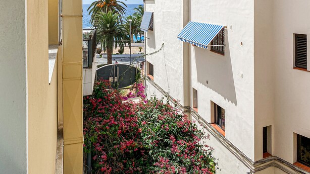 Une ruelle étroite d’où l’on aperçoit la mer, un palmier et un bâtiment surmonté d’une marquise à rayures bleues.