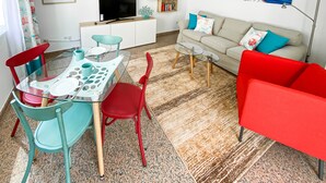 Un salon avec un canapé, des fauteuils rouges et turquoise, une table en verre, un téléviseur et une bibliothèque.
