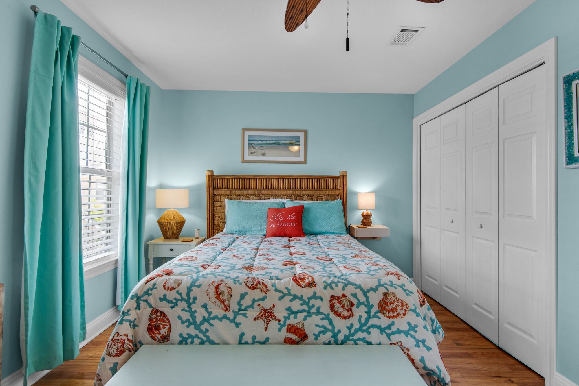 A bedroom with a bed, two bedside tables, a wooden headboard, a window with curtains, a ceiling fan, and a wardrobe.