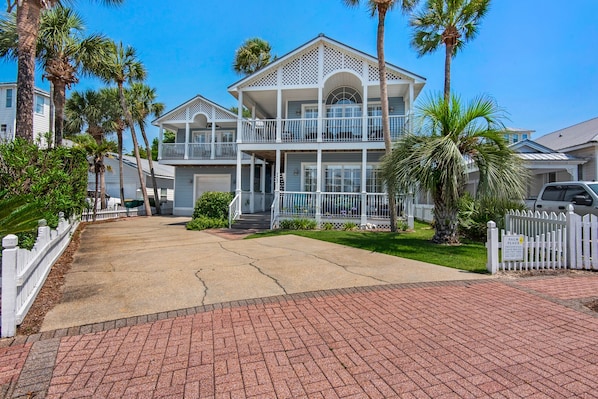 A two-story house with a balcony, surrounded by palm trees and a white picket fence.