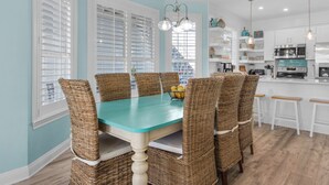 A dining area with a turquoise table and wicker chairs, a kitchen with white cabinets, and a hanging light fixture. - Palm Place, Spacious beach house steps to the powdery sand and emerald water. (Destin)