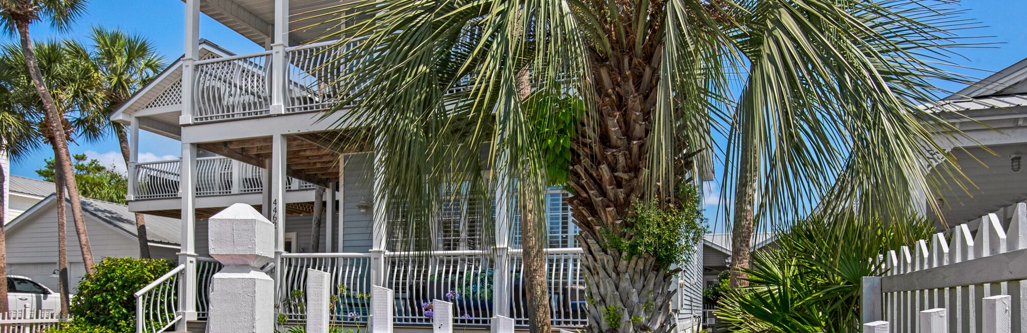 A multi-story building with balconies, surrounded by palm trees and a white fence.