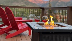 A deck with two red Adirondack chairs, a black fire pit with flames, and a glass of red wine.