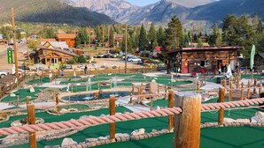 A miniature golf course with a wooden clubhouse, surrounded by mountains and a small town.