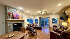 A spacious living room with a stone fireplace, leather sofas, a wooden dining table, and a ceiling fan.