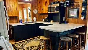 Une cuisine avec des meubles en bois, un plan de travail en marbre, un évier, une cuisinière, un four à micro-ondes et une horloge. Il y a des tabourets et un tapis.