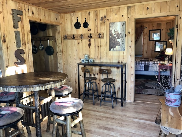 A rustic wooden interior with a round wooden table, four stools, and a bar area.