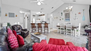 A modern living room with a leather couch, red cushions, a coffee table, and a dining area with white chairs and a wooden table. - Sandpiper Unit 4209/Beach Service/Gated Cmty/Reserved Parking/Many Amenities!!!! (Destin)