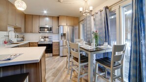 Cozinha moderna com eletrodomésticos de aço inoxidável, sala de jantar com mesa posta para dois e uma janela grande com cortinas azuis.
