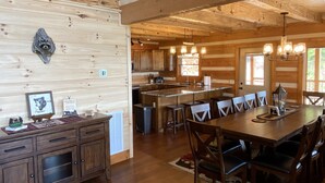A spacious wooden cabin interior with a large dining table, multiple chairs, a kitchen area, and a ceiling with exposed beams.