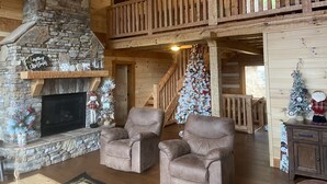 A cozy cabin interior with a stone fireplace, leather armchairs, a Christmas tree, and a wooden balcony.