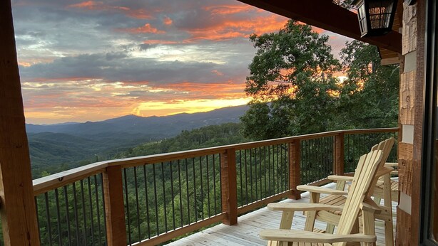 A wooden deck with Adirondack chairs overlooking a mountainous landscape during sunset.