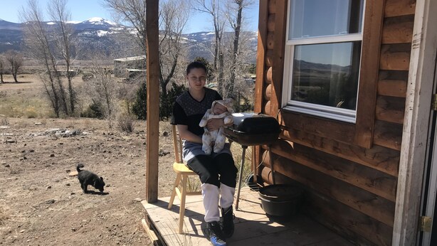 Une personne assise sur une chaise en bois, à l’extérieur d’un chalet, avec vue sur un paysage montagneux.