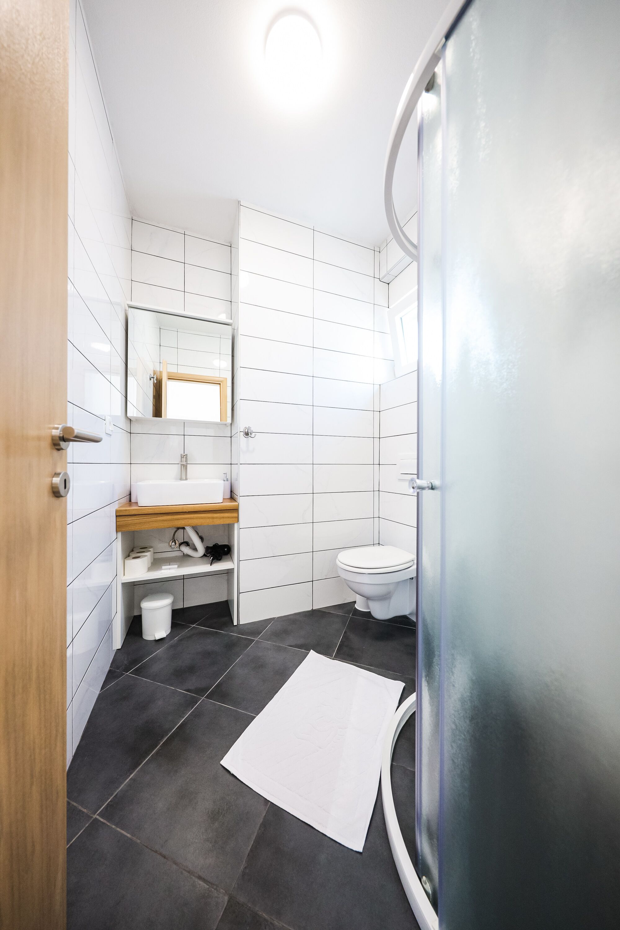 A modern bathroom with white tiles, a wooden vanity, a white sink, a toilet, and a glass shower enclosure.