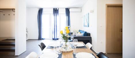 A dining area with a wooden table set for four, a vase with yellow flowers, and a view of a living room with a sofa and curtains.