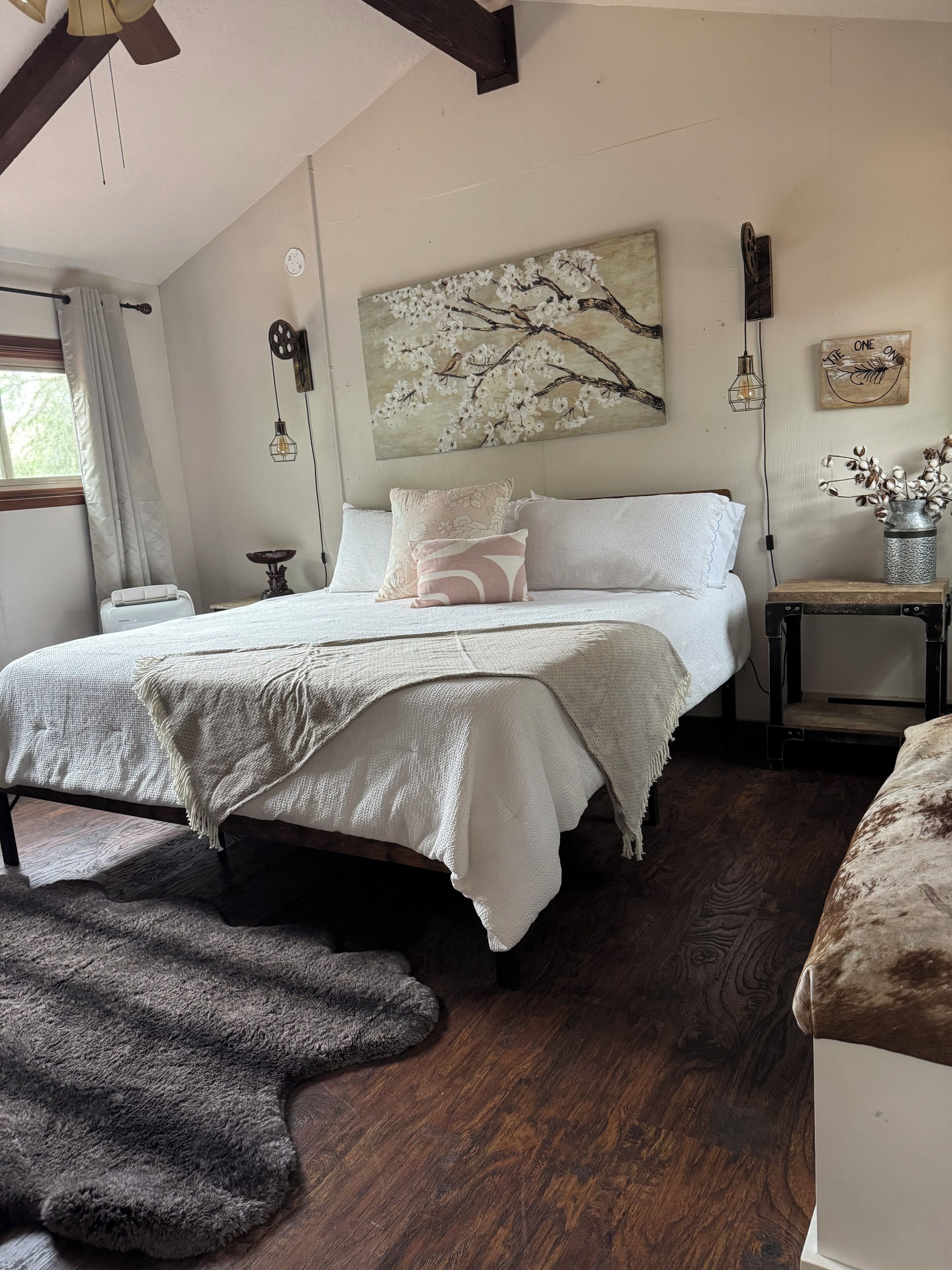 A bedroom with a bed, a window with curtains, a painting of a tree, a wall clock, and a small table with a vase.