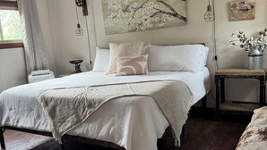 Um quarto com uma cama, uma janela com cortinas, um quadro de uma árvore, um relógio de parede e uma pequena mesa com um vaso.
