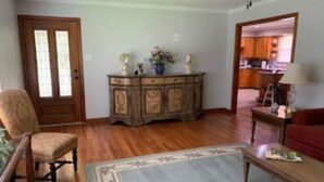 Un salon avec un plancher en bois, un tapis à motifs, un meuble en bois orné d’objets décoratifs, un fauteuil et un canapé rouge.