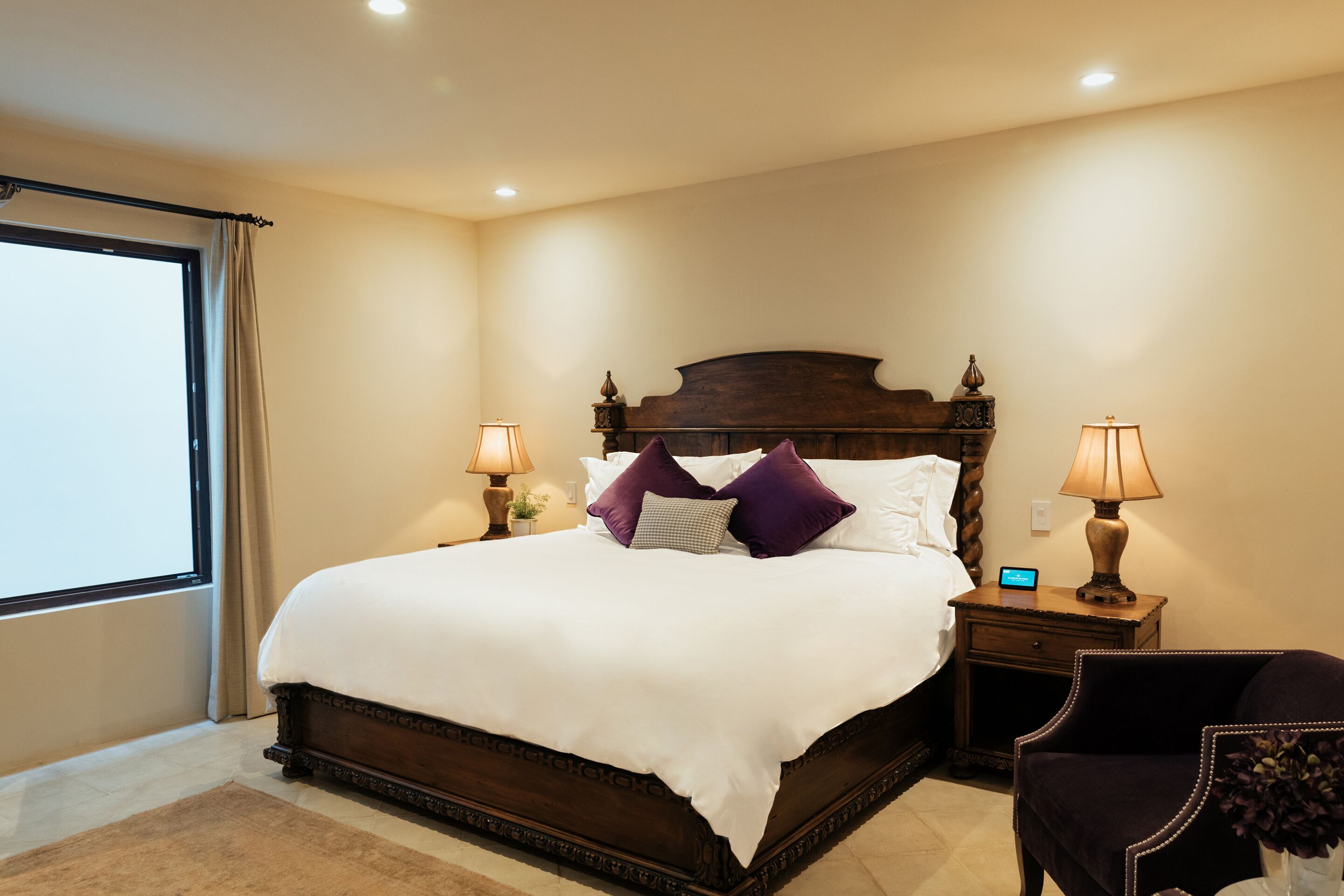 A neatly made bed with a wooden headboard, two bedside tables with lamps, a dark armchair, and a large window with curtains.