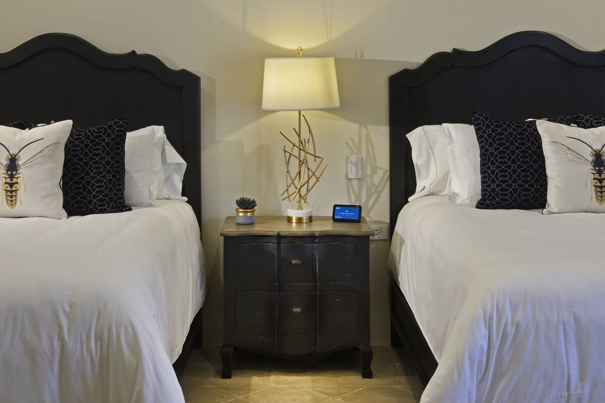Et hotelværelse med to senge, begge med sengegavl, hvidt sengetøj og dekorative puder med biedesign. Der er et natbord med lampe, vækkeur og en lille plante.