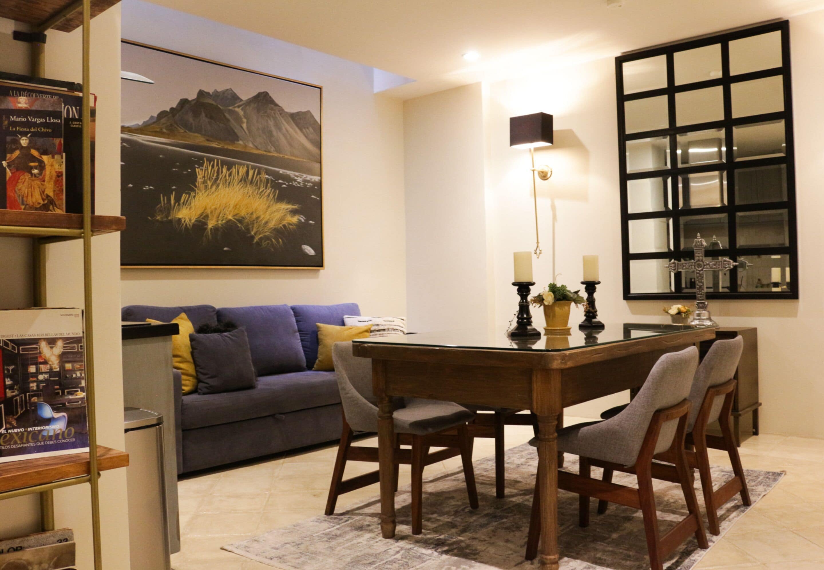 A dining area with a wooden table, four chairs, a candle holder, and a painting of mountains on the wall.