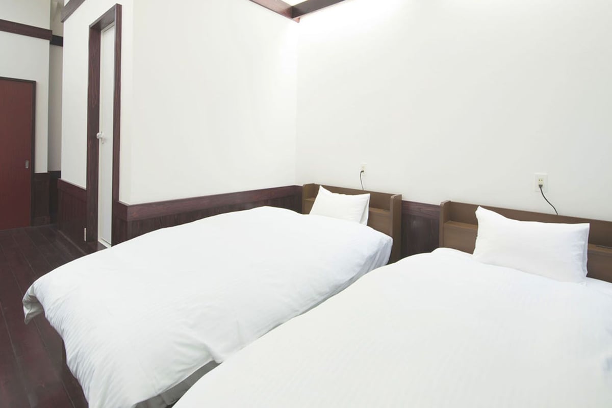 Habitació d'hotel amb dos llits individuals, ambres de fusta, paret blanca i una porta.