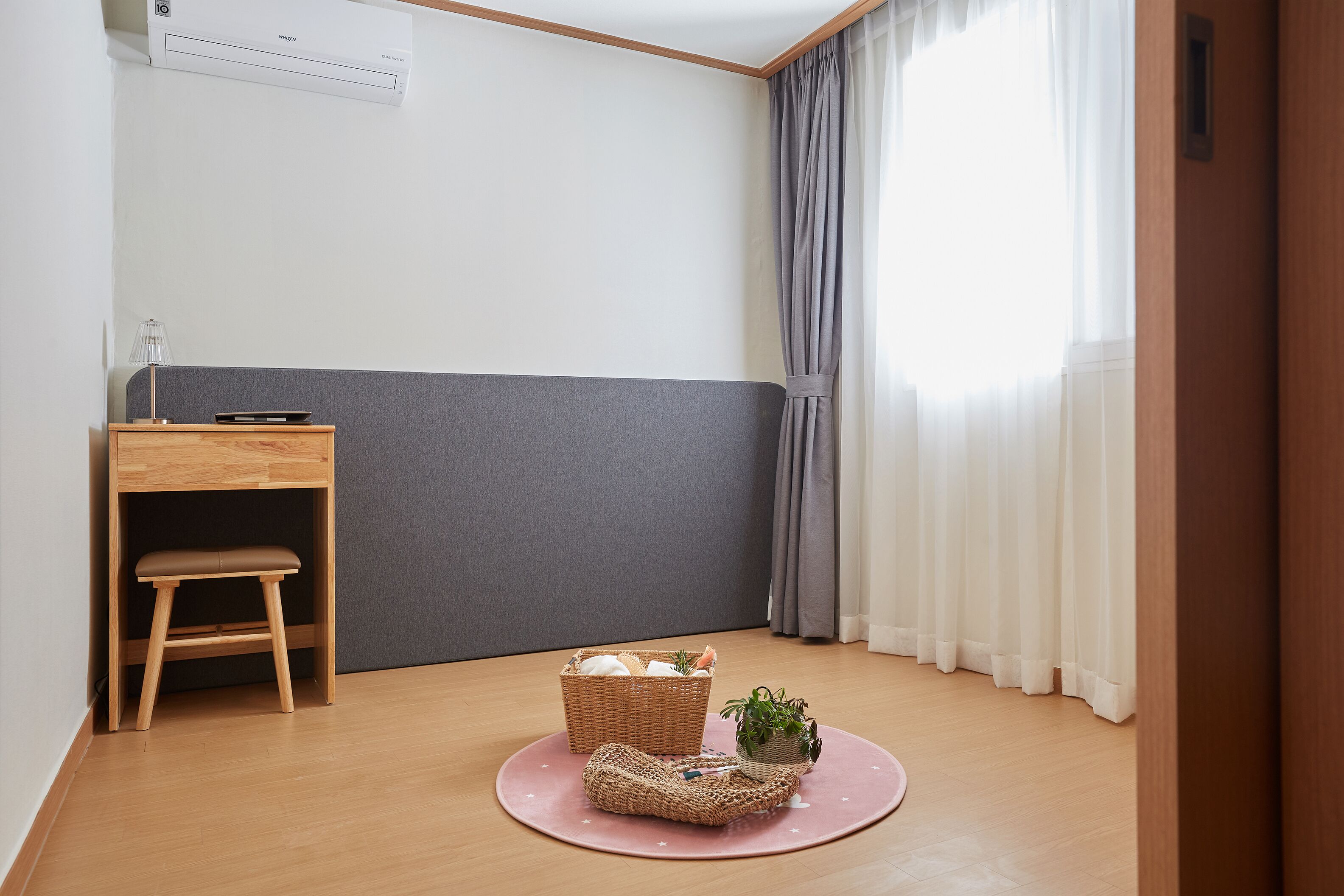 Een kamer met een houten bureau, een kruk, een rieten mand en een kleine plant op een roze mat.