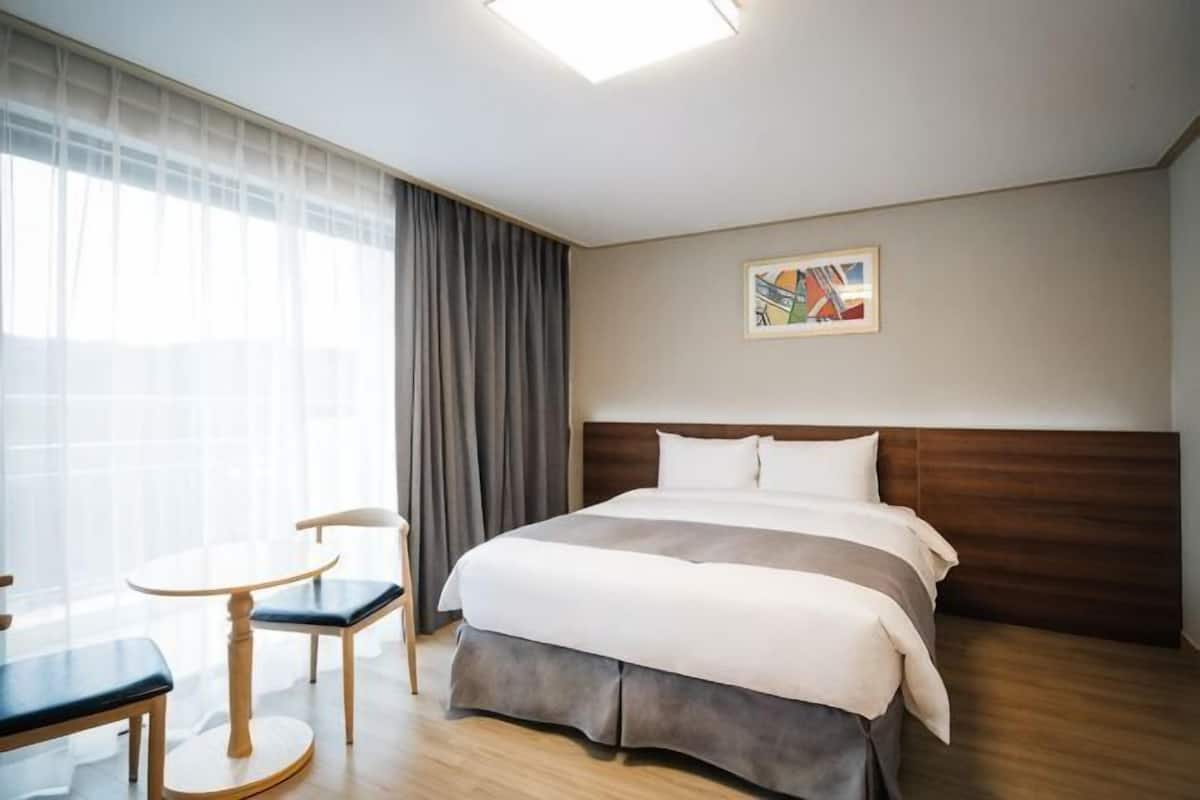 Ett hotellrum med en säng, ett litet bord med stolar, ett fönster med gardiner och en tavla på väggen.