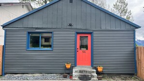 Un petit bâtiment d’un seul étage, avec une porte rouge et des bordures bleues, une fenêtre et deux plantes en pot placées sur les marches de l’entrée.