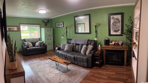 Un salon avec un canapé en cuir marron foncé, une table basse en bois et un tapis à motifs. Un mur vert est orné d’une tête d’animal fixée au mur et d’un tableau encadré. On peut également apercevoir un meuble en bois et une petite table avec une lampe.