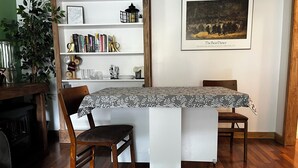 Un coin repas avec une table recouverte d’une nappe à motifs floraux, entourée de chaises en bois. On aperçoit, à l’arrière-plan, une bibliothèque remplie de livres et d’objets décoratifs.