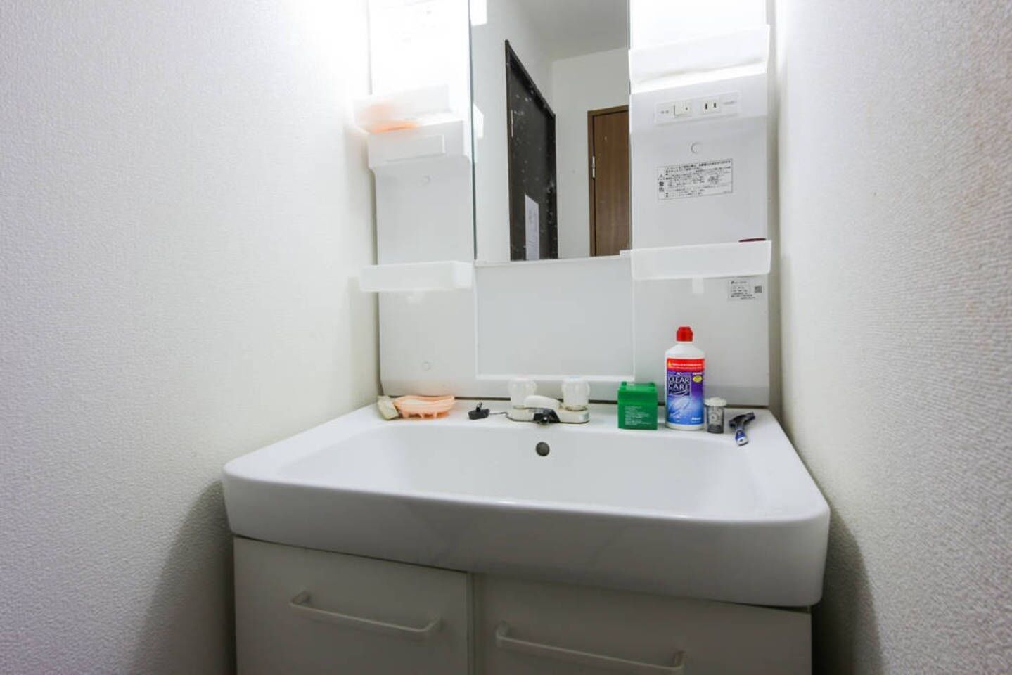 A white bathroom sink with a mirror above it, a small shelf with toiletries, and a door in the background.