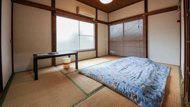 A traditional Japanese room with tatami flooring, a bed with a floral bedspread, a small table, and a hanging light fixture. - Nishiogi House Maipenrai (Tokyo)
