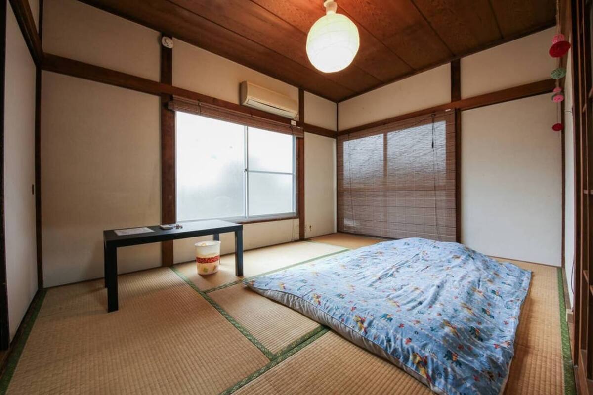 A traditional Japanese room with tatami flooring, a bed with a floral bedspread, a small table, and a hanging light fixture.