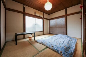 Une pièce de style japonais traditionnel, avec un sol recouvert de tatamis, un lit avec une couvre-lit à motifs floraux, une petite table et un luminaire suspendu.