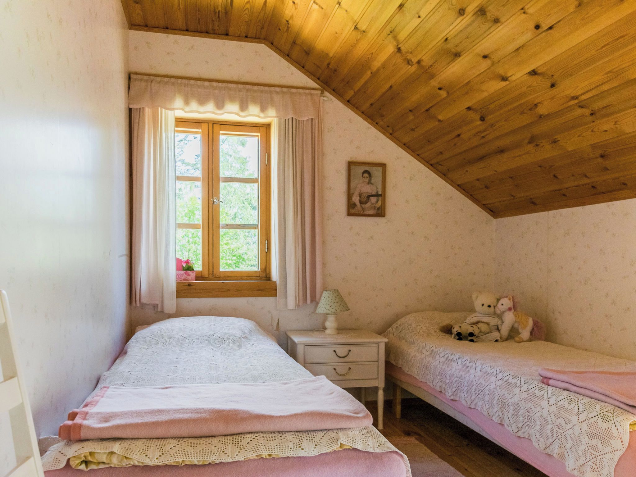 Ein Schlafzimmer mit schrägem Dach, einem Fenster mit Vorhängen, einem Nachttisch mit Lampe und zwei Betten.