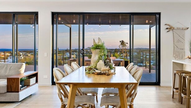 A spacious dining area with a long wooden table and wicker chairs, large windows offering a city view, and a macrame wall hanging. - Hedge Apartments by CLLIX (Buddina)
