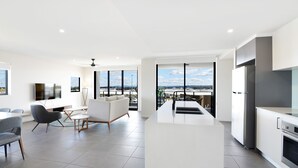 A modern kitchen with a large island, a dining area with a sofa and chairs, and large windows offering a city view. - Hedge Apartments by CLLIX (Buddina)
