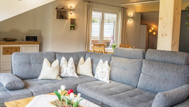 Ein modernes Wohnzimmer mit grauem Sofa, dekorativen Kissen, einem Holztisch und einem Essbereich im Hintergrund.